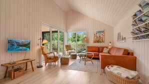 A cozy living room with a wooden floor, a large window, a television, a sofa, and a bookshelf. - Traumhaus Römö für 6 Personen. Unesco Wattenmeer (Rømø)