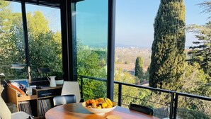 A dining area with a wooden table and chairs, a bowl of fruit, and a view of a pool and greenery through large windows. - Villa D'architecte Avec Piscine au Coeur D'un Parc (Villeneuve-lès-Avignon)