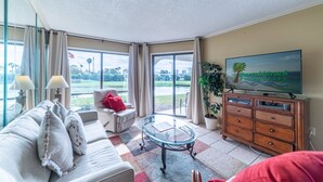 A living room with a sofa, a glass coffee table, a TV on a wooden cabinet, and a view of a golf course through large windows.