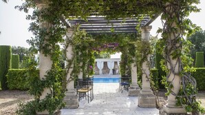 Un área de estar al aire libre con techo, columnas de piedra, piscina y setos bien cuidados.