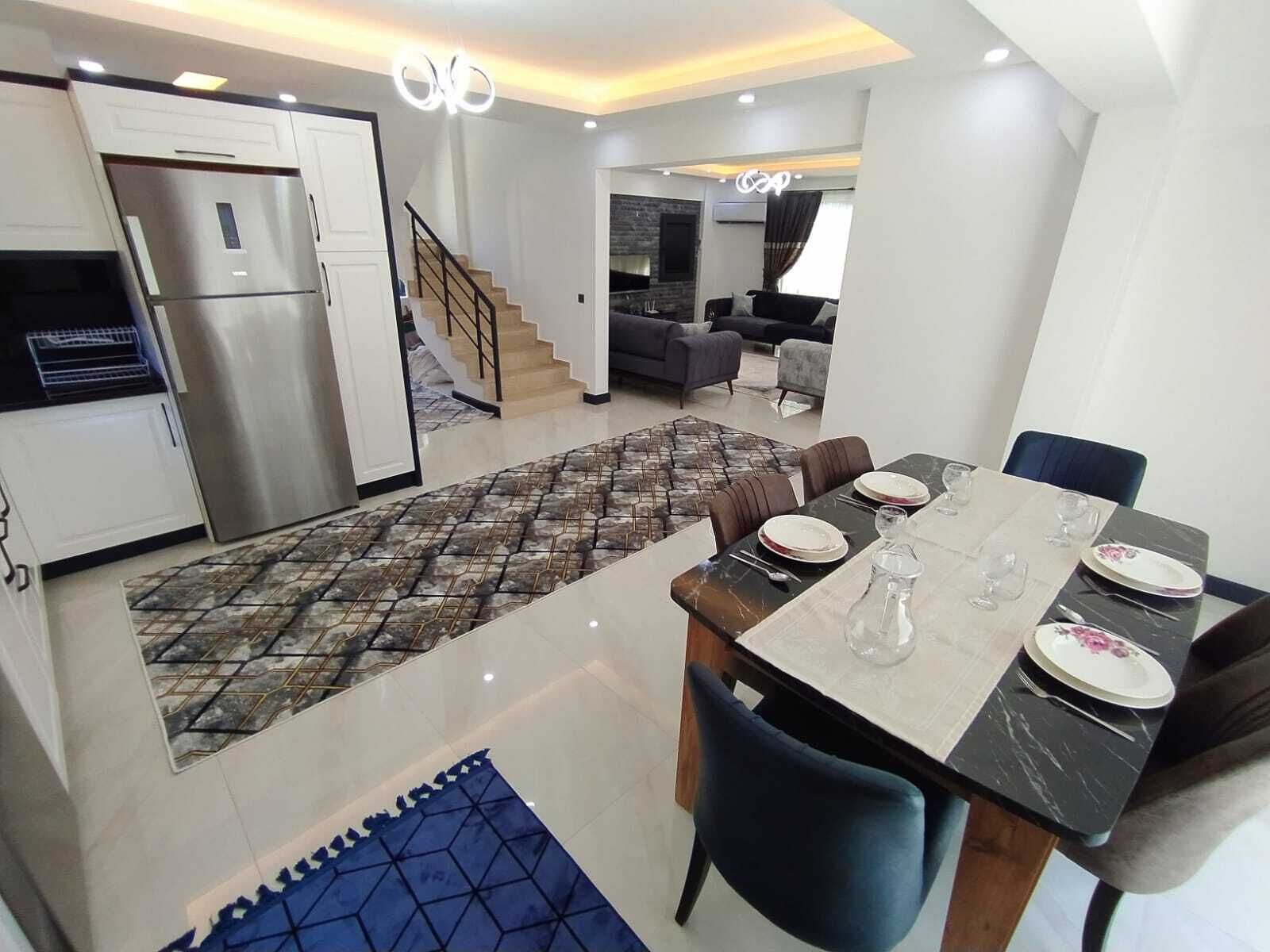 A modern kitchen with a built-in refrigerator, a dining area with a marble table, and a staircase leading to a living room.