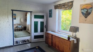 Une pièce avec un meuble en bois, une fenêtre donnant sur un paysage verdoyant, une porte avec des volets verts et une chaise à motifs.