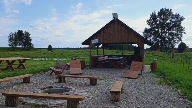 Un pavillon rustique en bois, doté de bancs et d’un foyer, entouré d’un champ herbeux et d’arbres.