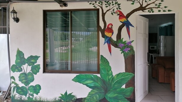 A room with a large window, a mural of parrots and foliage, and a visible kitchen area.