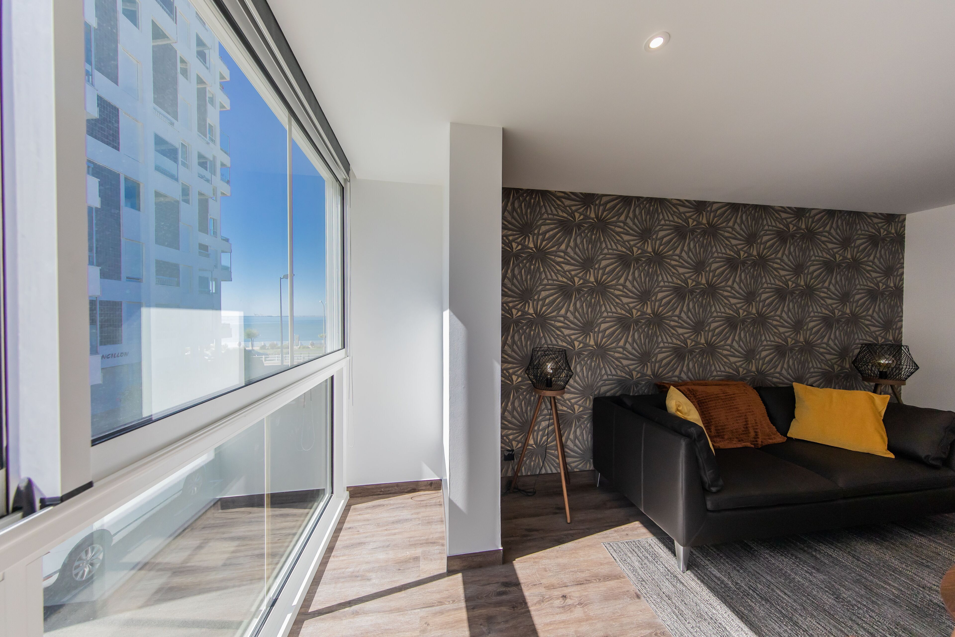 Een moderne woonkamer met een zwarte bank, behang met een patroon en een groot raam met uitzicht op een gebouw.