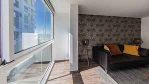 A modern living room with a black sofa, patterned wallpaper, and a large window offering a view of a building. - Le Foncillon 4* Apartment at the foot of the beach (Royan)