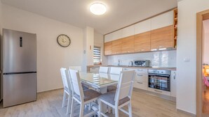 Eine moderne Küche mit einem Edelstahl-Kühlschrank, einem Esstisch mit Glasplatte und weißen Stühlen, Holzschränken, einer Mikrowelle und einem Backofen.