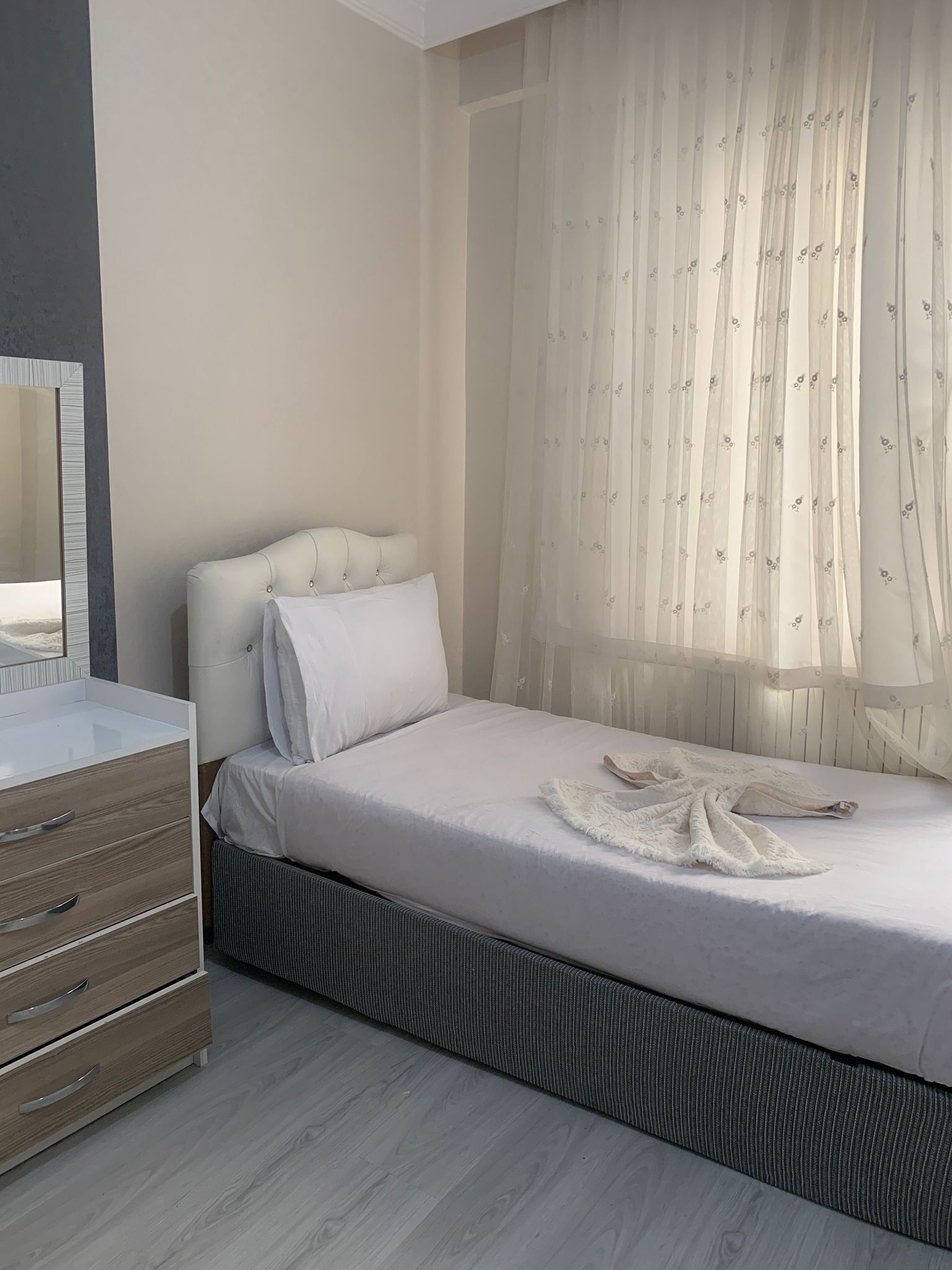 Een slaapkamer met een bed, een dressoir en een raam met dunne gordijnen.