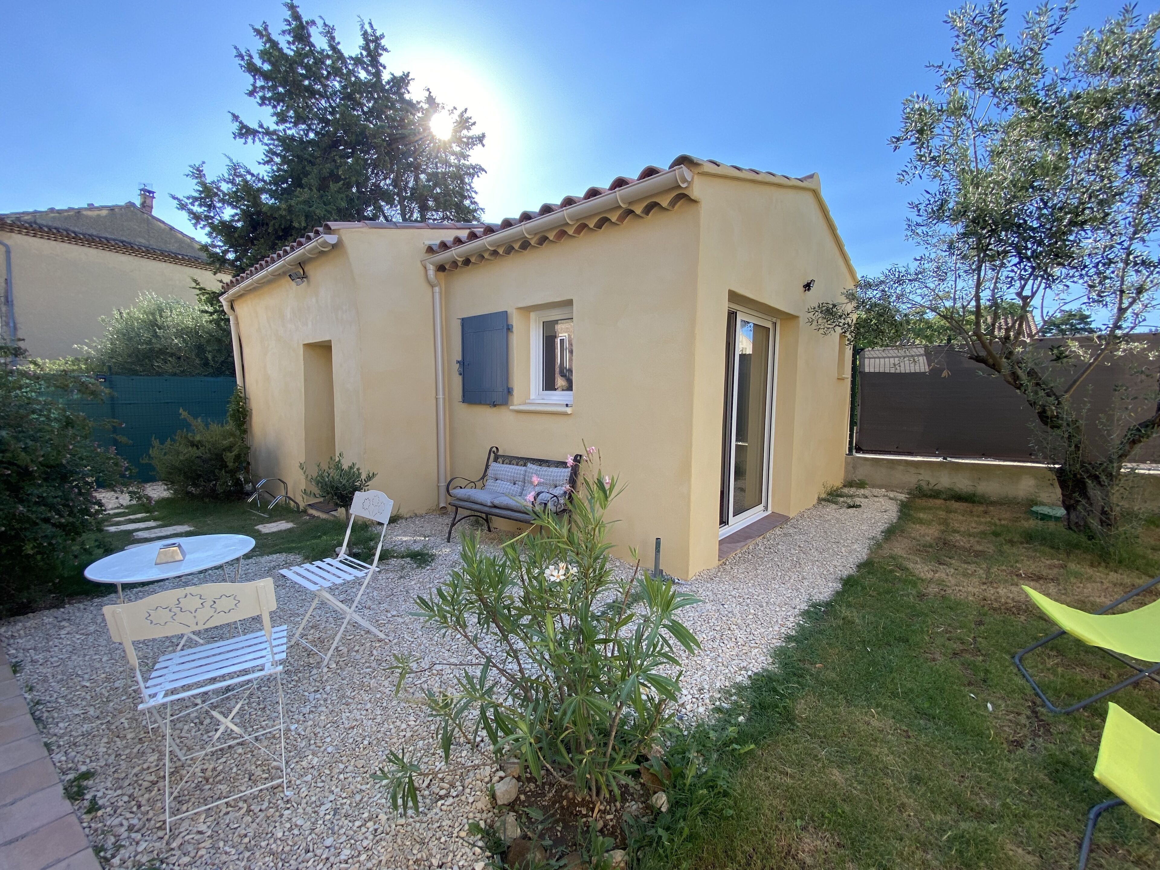 A small, single-story building with a tiled roof, a patio area with a table and chairs, and a garden with various plants.