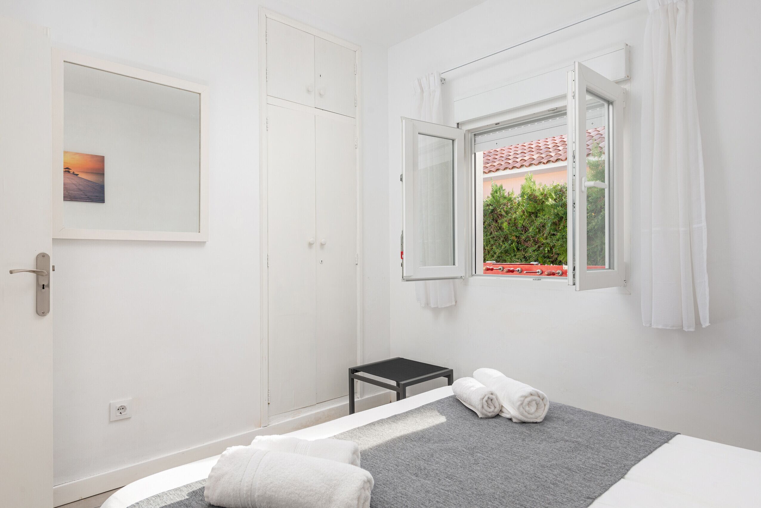 Hotellihuone, jossa on sänky, pieni pöytä, ikkuna, jonka sälekaihtimet ovat auki, peili ja taulu seinällä.