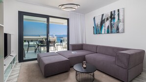 Un salon moderne avec un canapé d’angle, une petite table ronde et une vue sur la mer grâce à de grandes portes vitrées.