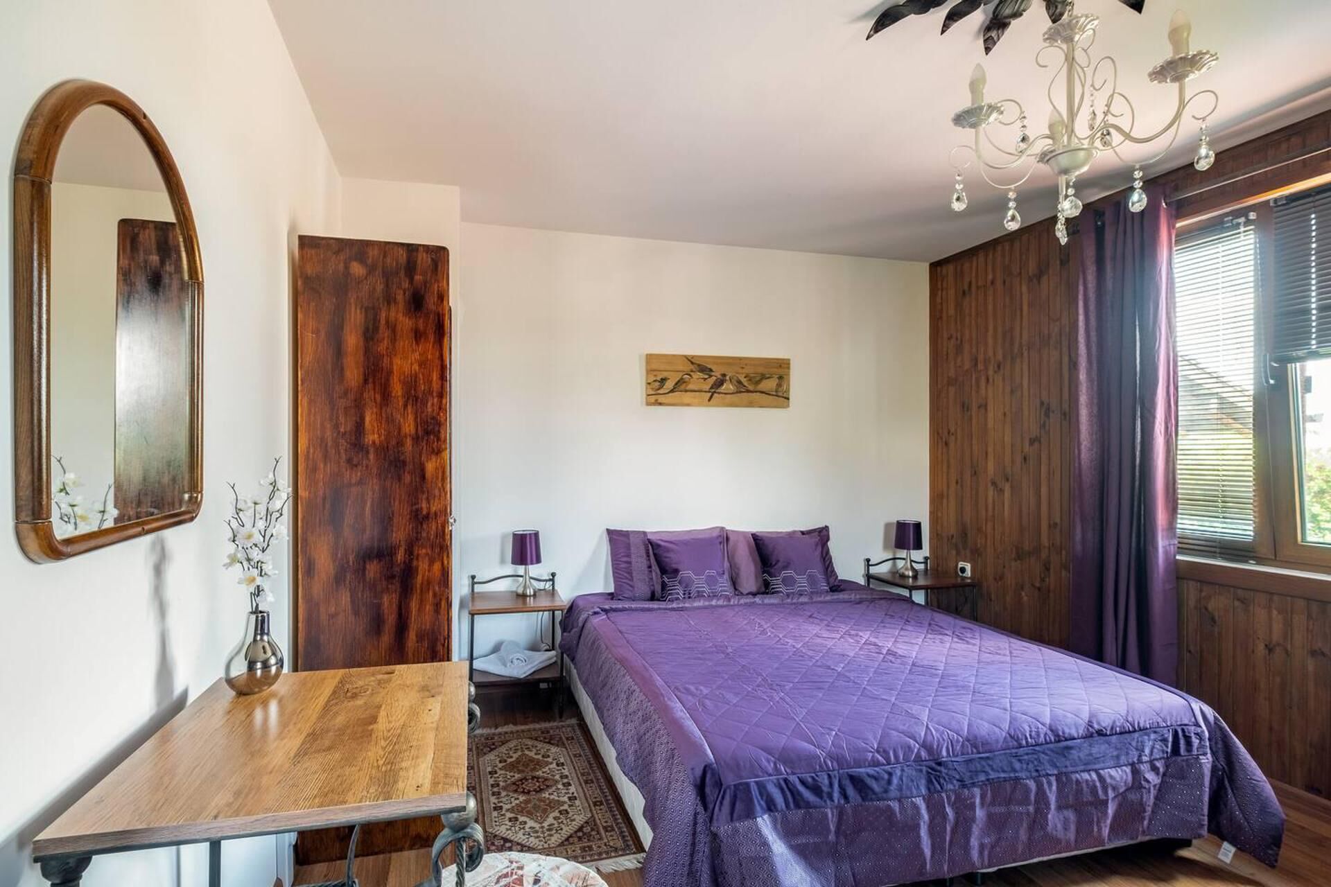 A bedroom with a bed, a wooden nightstand, a table, a mirror, a chandelier, and a window with curtains.