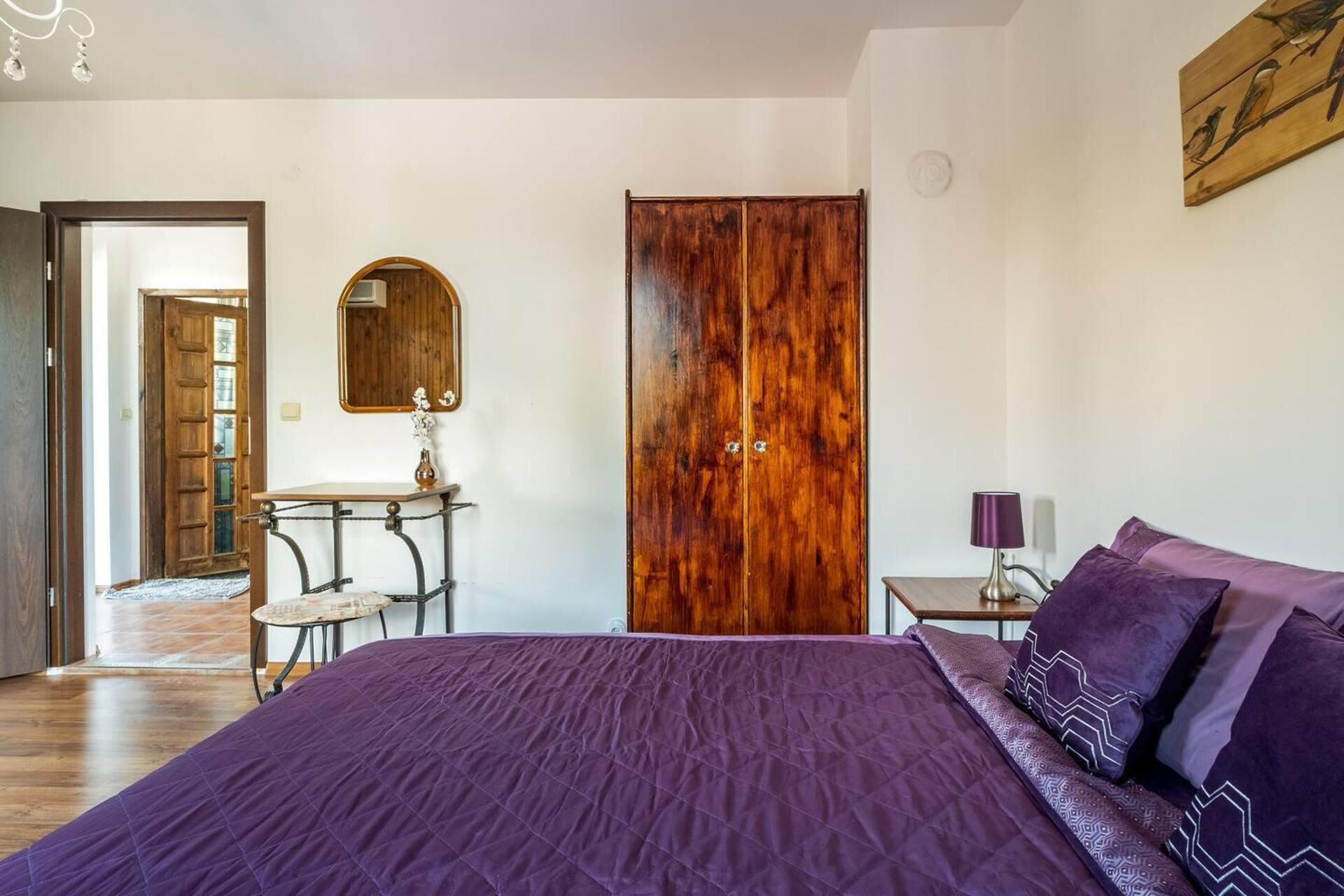 A bedroom with a large bed, a wooden wardrobe, a small table with a mirror, and a bedside lamp.