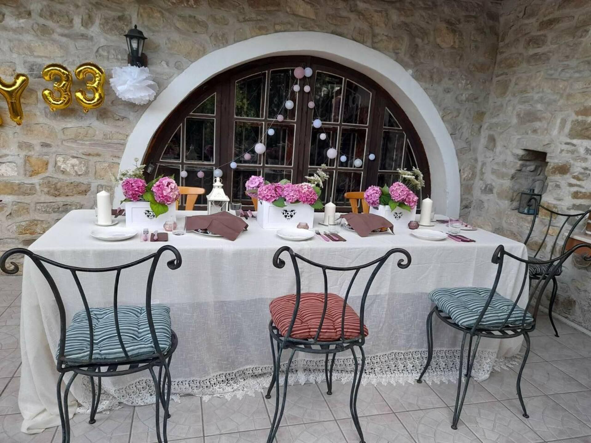 A table set for a meal with a stone wall backdrop, a large arched window, and decorative balloons.