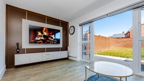 A modern living room with a flat-screen TV, a glass coffee table, and a view of a brick wall and greenery outside. - My Chester Luxury 3bedroom Home (Colchester)