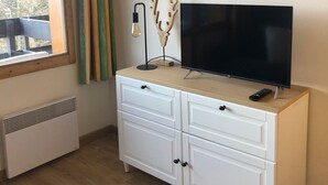 A room with a TV on a wooden cabinet, a window with curtains, and a radiator. - Studio 4 Personnes Proche des Pistes (La Plagne-Tarentaise)