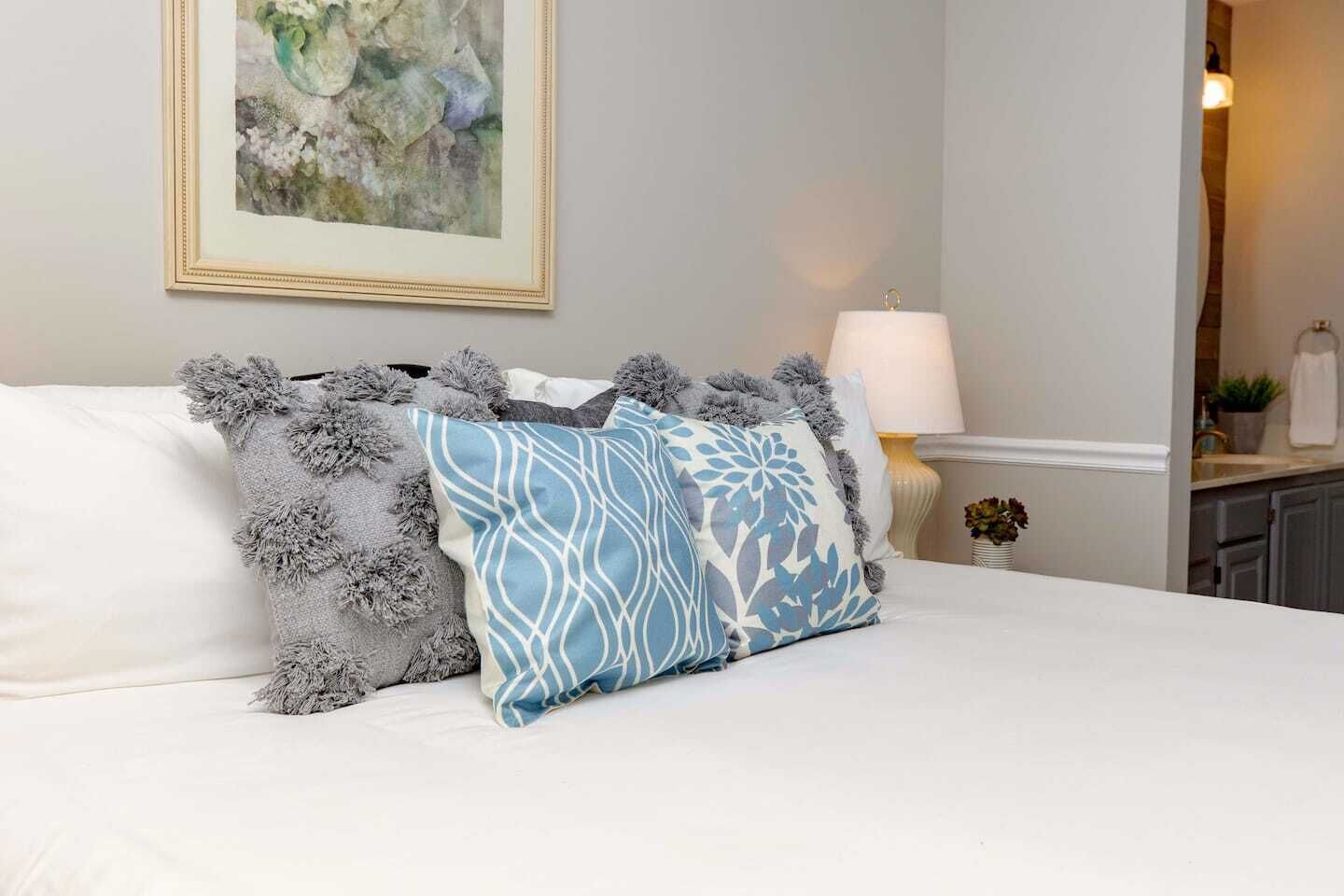 Una cama con ropa de cama blanca y dos almohadones decorativos, uno con estampado azul y el otro con textura gris y esponjosa.