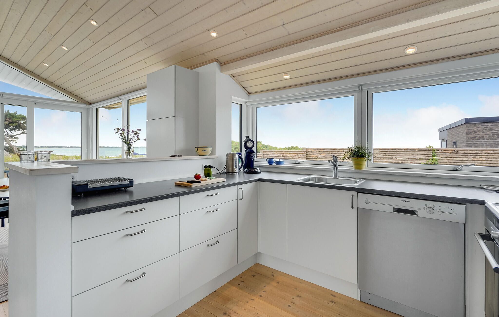 A modern kitchen with white cabinetry, a wooden ceiling, and large windows offering a view of the beach.