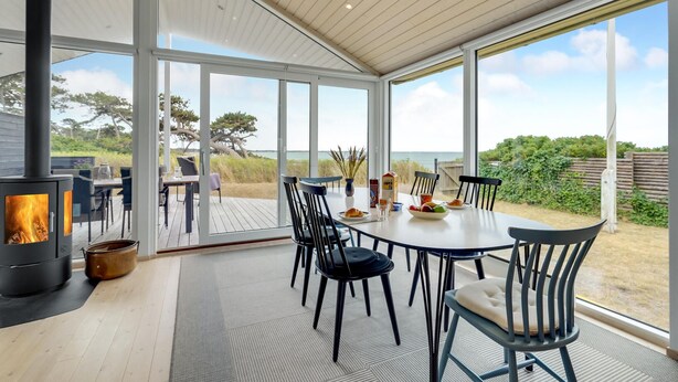 A modern outdoor dining area with a large table and chairs, a wood-burning stove, and a view of the beach and dunes.