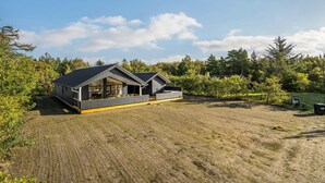 A modern cabin with a large deck and a covered porch, surrounded by a forested area and a dirt field.