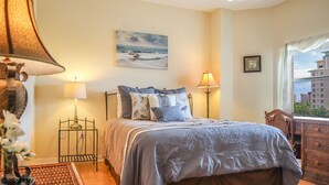 A bedroom with a bed, two bedside lamps, a ceiling fan, a window with a view of buildings, and a painting on the wall. - GRAND EXECUTIVE 3B/3B WATERSIDE 246 ISLAND CONDO (Fort Myers Beach)