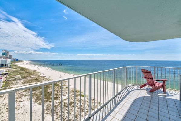 A balcony with a red Adirondack chair overlooking a beach with a pier and buildings in the background.