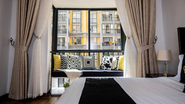 Ein Schlafzimmer mit einem großen Bett, einem Fenster mit Vorhängen, Blick auf ein Gebäude und einer Lampe auf einem Nachttisch.