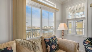A living room with a beige sofa, floral cushions, a lamp, and large windows with a view of a cityscape. - Sunset Island-Fountain Dr 37 #5A-Bayside 67th St, WIFI, Pools, Elev, W/D, AC (Ocean City)