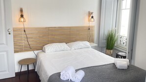 Siististi pedattu sänky, jossa on valkoiset lakanat, harmaa heittopeitto ja rullalle taitetut pyyhkeet harmaalla jalkarahilla. Sängyn molemmilla puolilla on seinään kiinnitetyt valaisimet, ja ikkunan lähellä näkyy ruukussa oleva kasvi.