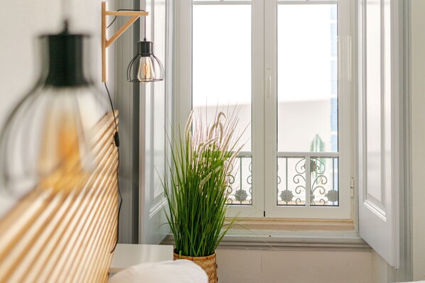 Ein Zimmer mit einem Bett, einer Hängelampe, einem Topfpflanzen und einem Fenster mit Blick auf einen Balkon.