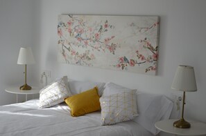 A neatly made bed with white and gold pillows, a white wall, and a floral artwork above it.