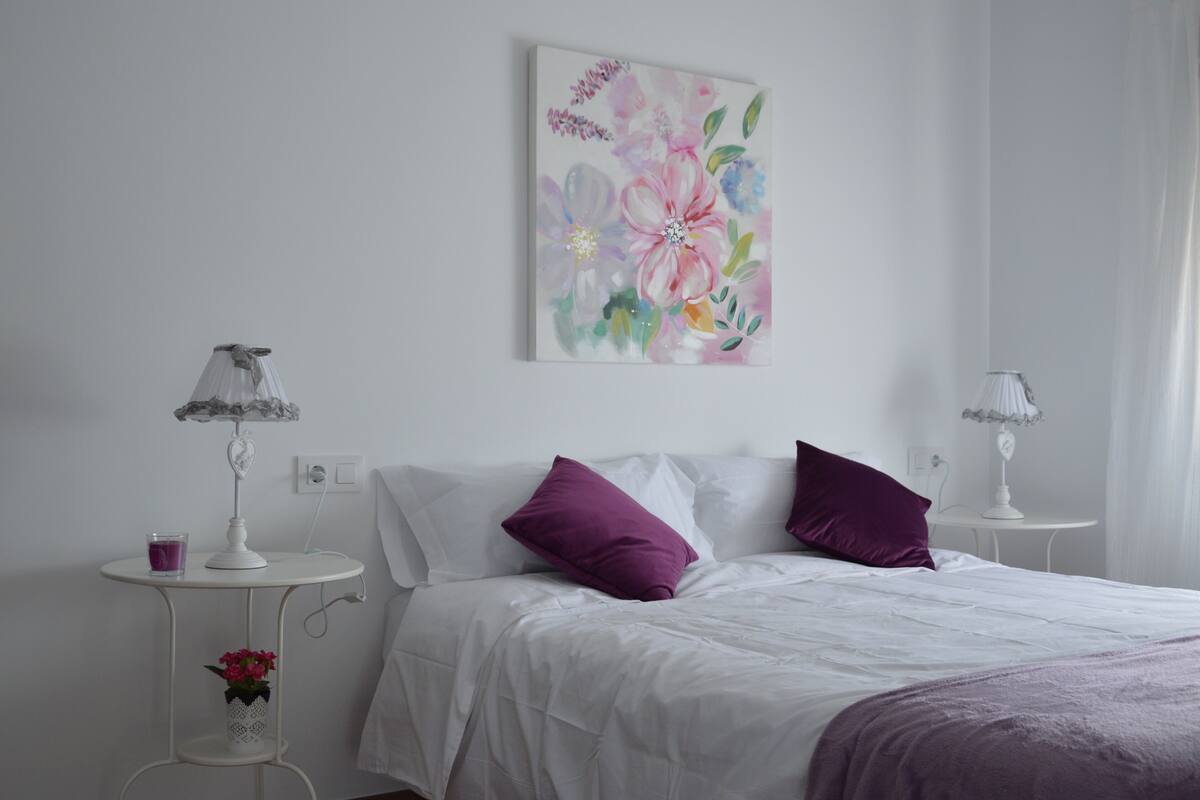 A bedroom with a bed, two nightstands, a wall-mounted lamp, a bedside table, and a floral painting.