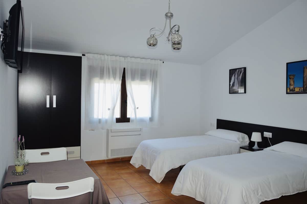 A hotel room with two single beds, a TV, a table with a chair, a window with curtains, and a chandelier.