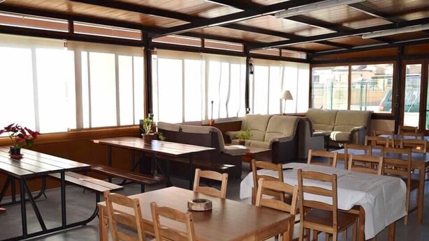 A dining area with wooden tables and chairs, a sofa, and large windows allowing natural light.