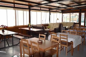 Uma sala de jantar com mesas e cadeiras de madeira, um sofá e grandes janelas que permitem a entrada de luz natural.