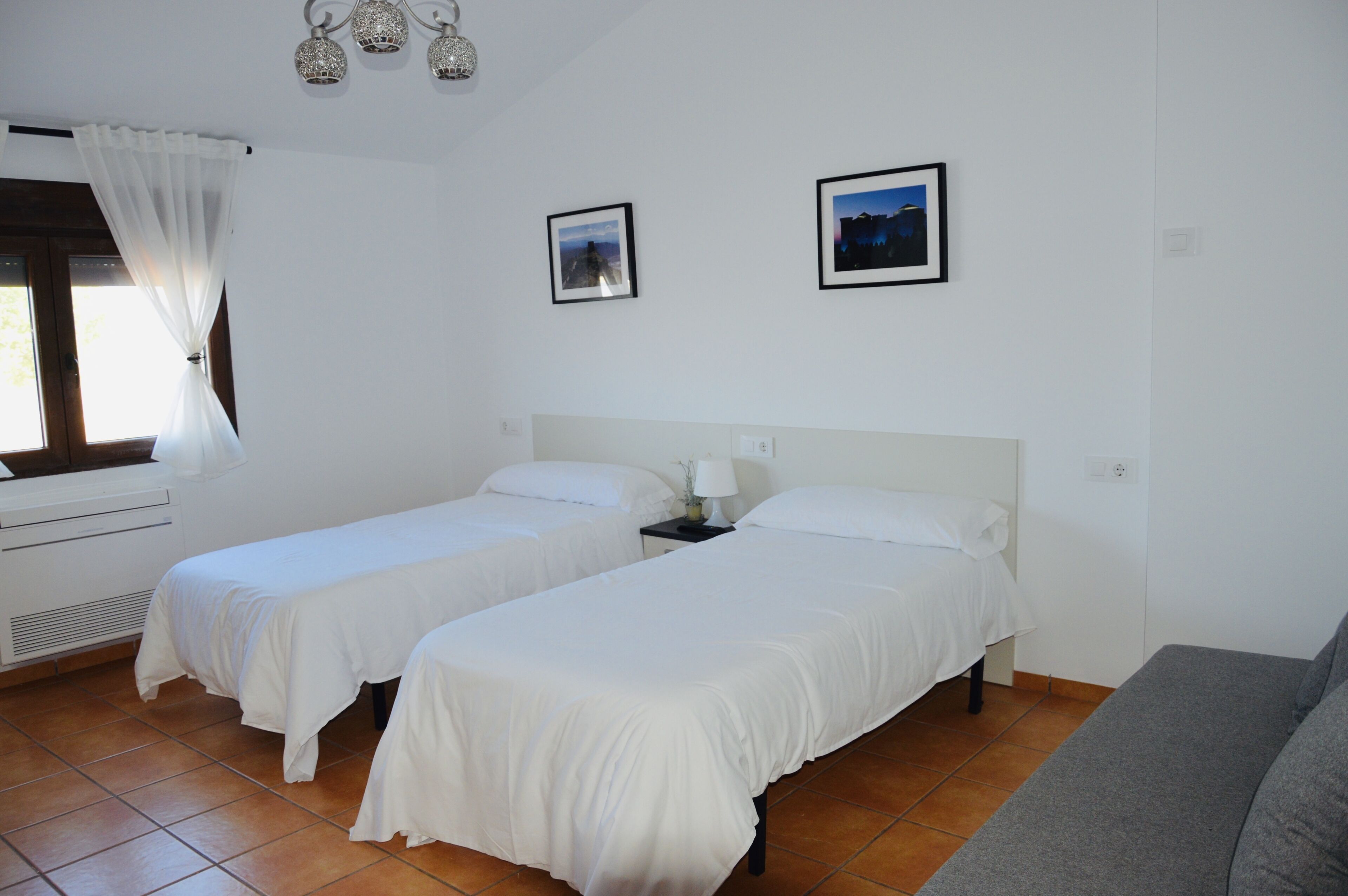 Une chambre avec deux lits simples, un canapé gris, une fenêtre avec des rideaux blancs et deux tableaux encadrés au mur.
