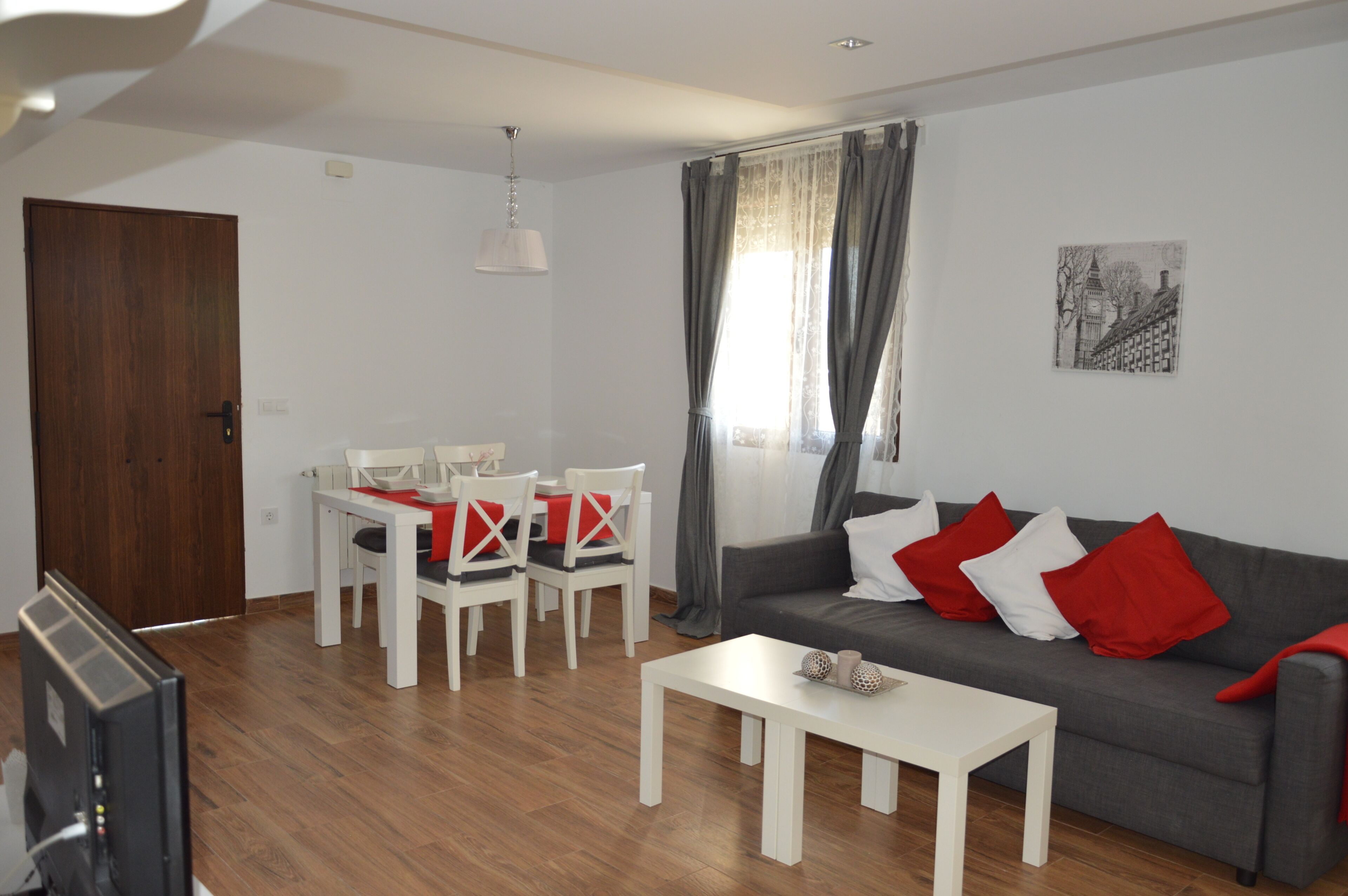 Una sala moderna con un sofá gris, una mesa de centro blanca y cojines rojo y blanco. Hay una mesa de comedor con sillas y un televisor en una esquina.