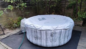Un bain à remous de forme ronde, doté d’une structure extérieure en bois et d’un couvercle, installé sur un tapis texturé à l’extérieur.