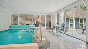 A swimming pool with a glass enclosure, a sun terrace with chairs, and a view of a thatched-roof building. - Stunning home in Blåvand with sauna (Blåvand)