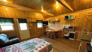 Een gezellig vakantiehuisje met houten inrichting, een bed met sprei, een eethoek met stoelen, een keuken met apparatuur en een raam met gordijnen.