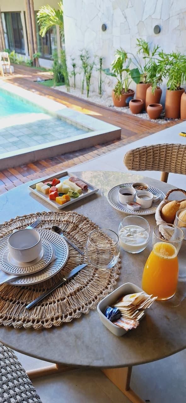 A dining table set with breakfast items, including fruits, bread, and beverages, overlooking a swimming pool and surrounded by potted plants.