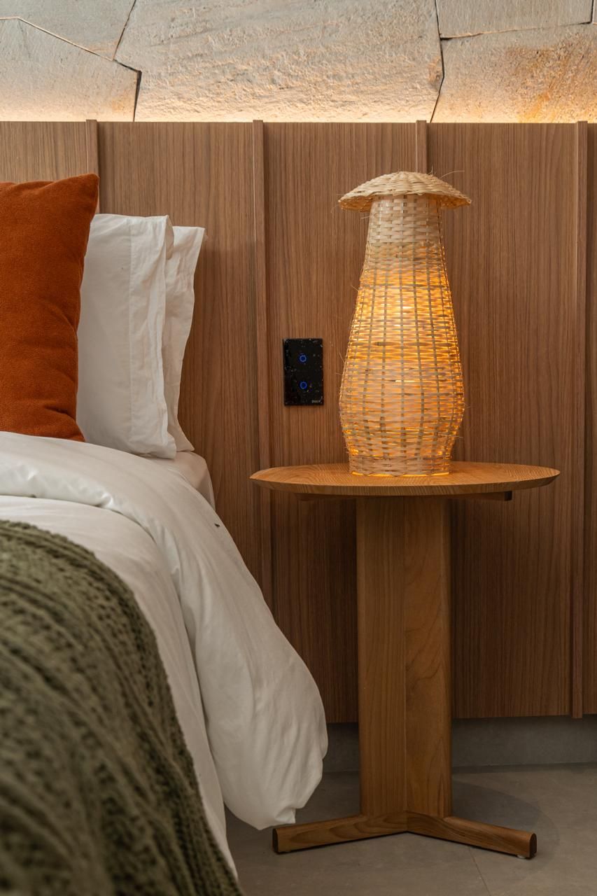 A bed with white and orange pillows, a woven lamp on a wooden table, and a control panel on the wall.