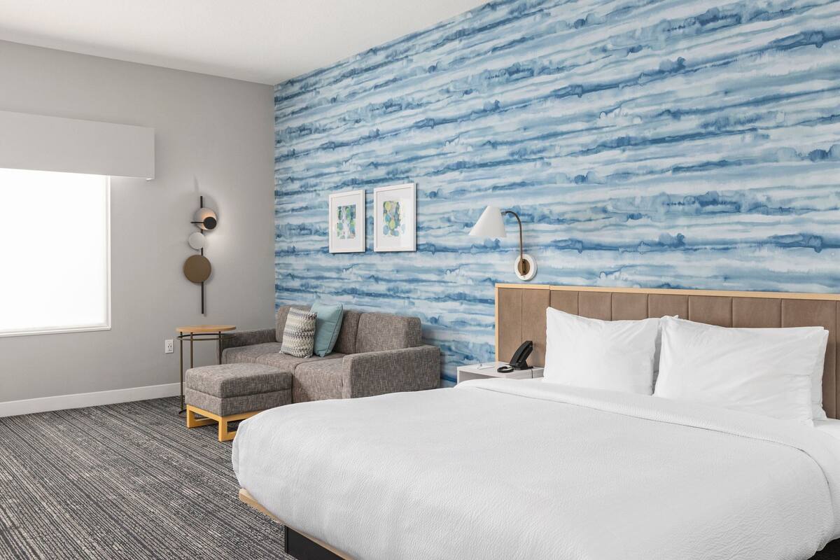 Ein Hotelzimmer mit einem großen Bett, einem grauen Sofa, einem kleinen Tisch und einer Wand mit einem blauen, abstrakten Muster.