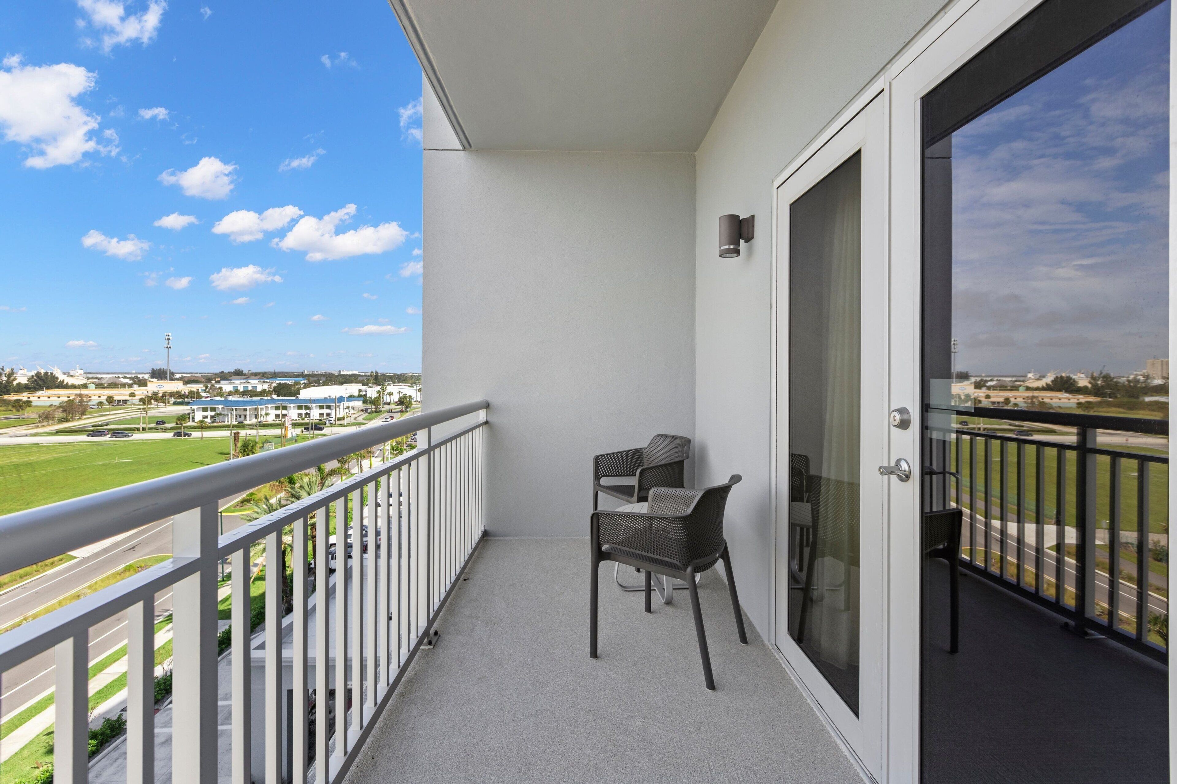A balcony with two chairs, a glass door, and a view of a green field and buildings.