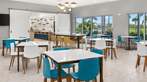 A modern dining area with white and blue chairs, a central bar, and large windows offering a view of palm trees and buildings.