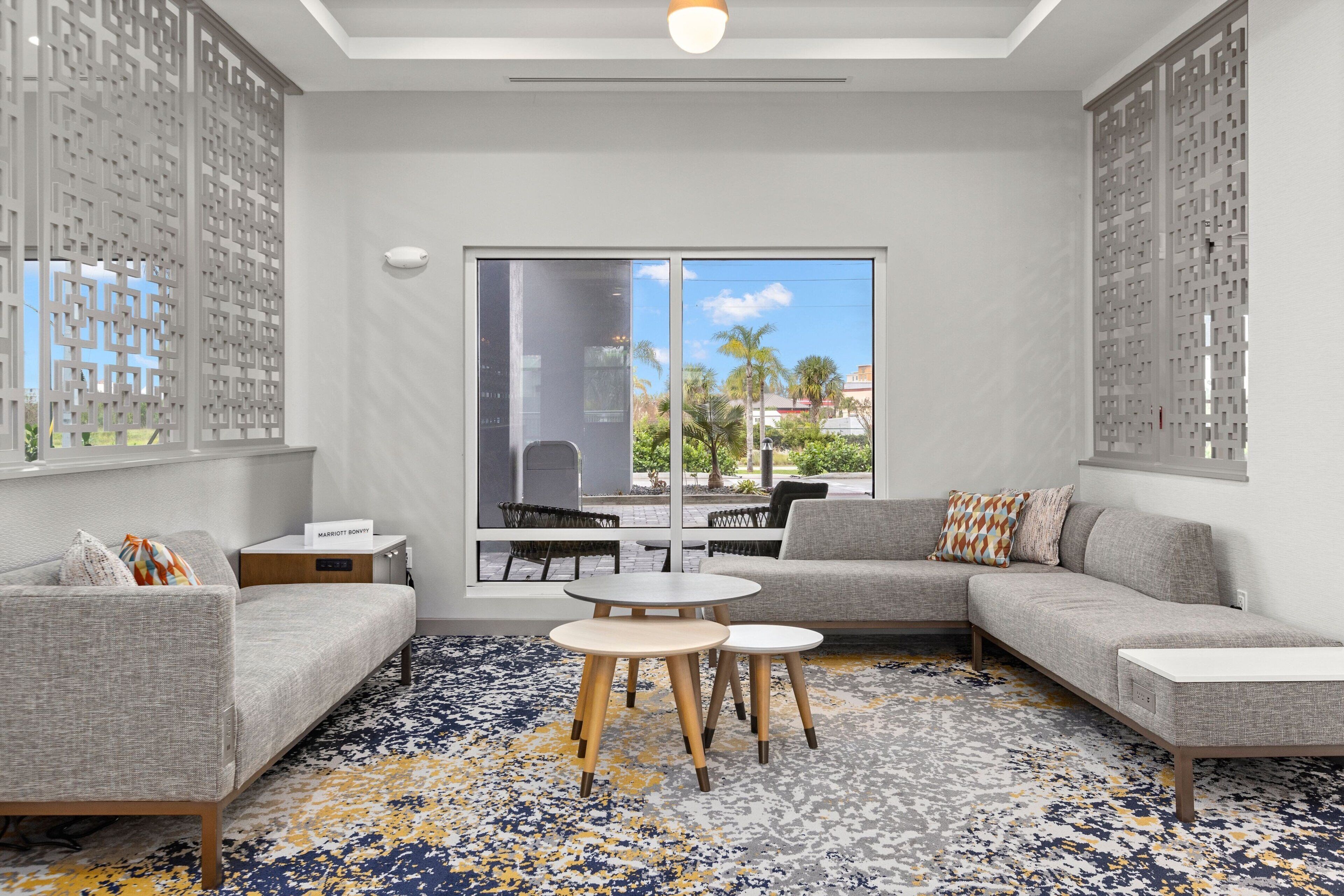 A modern living room with a grey sofa, two round tables, and a large window with a view of palm trees and buildings.