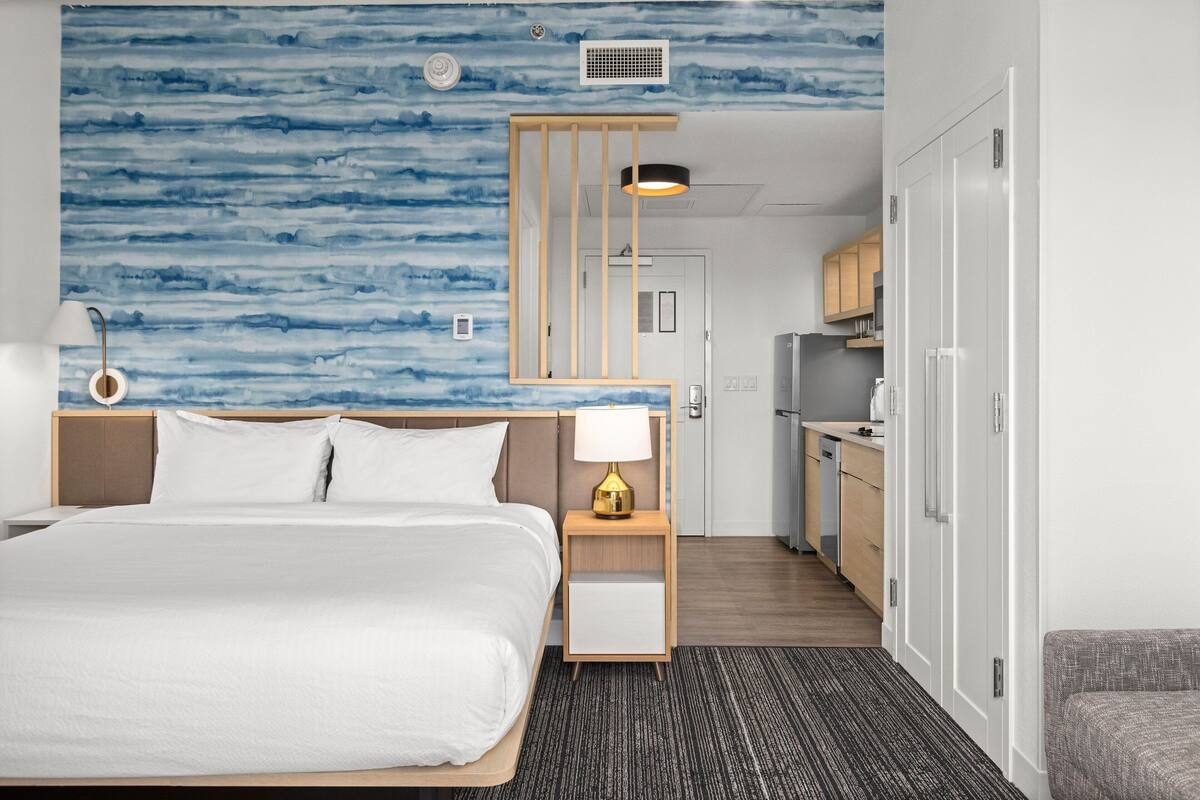 Ein Hotelzimmer mit Bett, Nachttisch, Lampe und Blick auf die Küche.