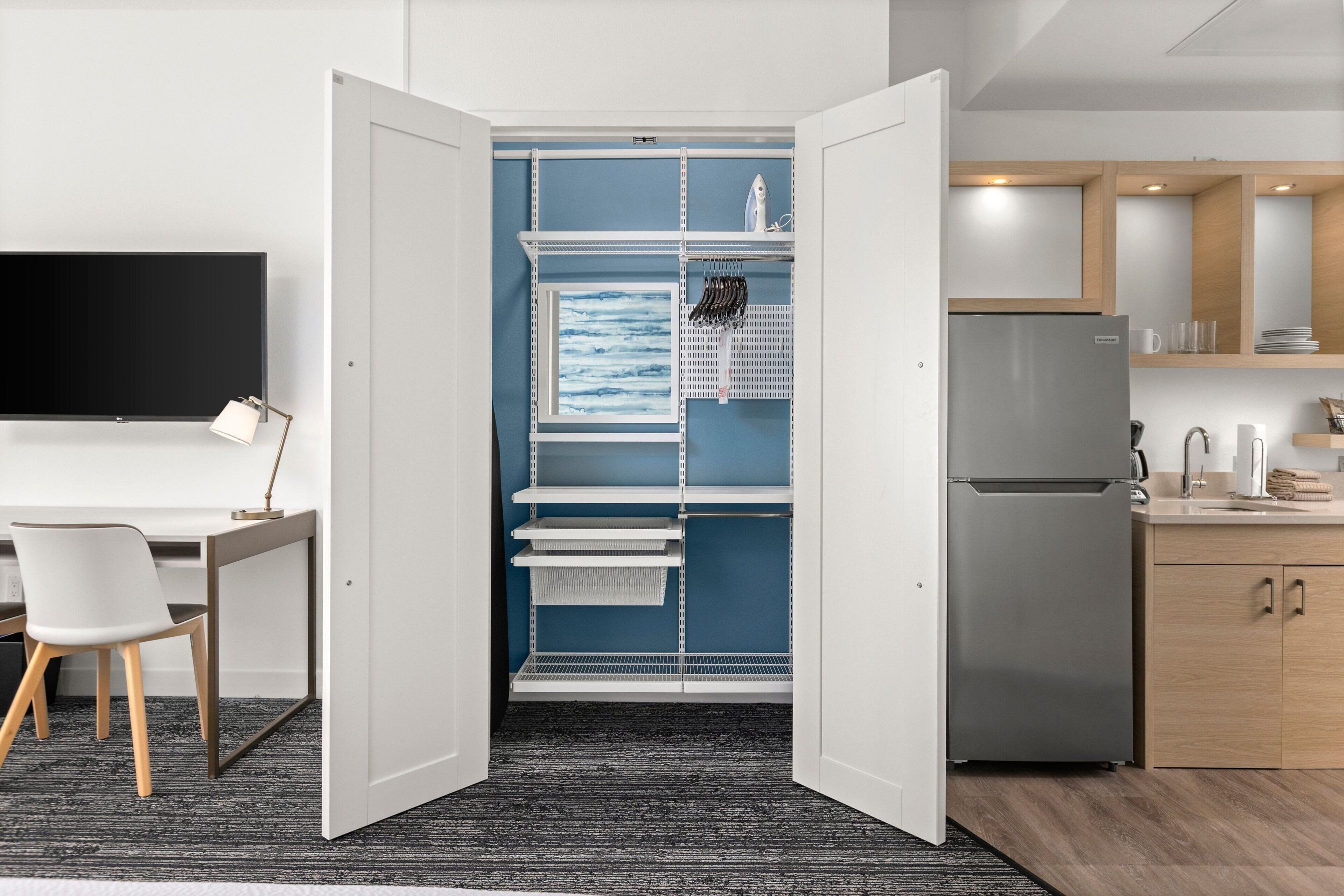 Quarto moderno com escrivaninha, cadeira e televisão integrada. Possui cozinha compacta com geladeira e armários.