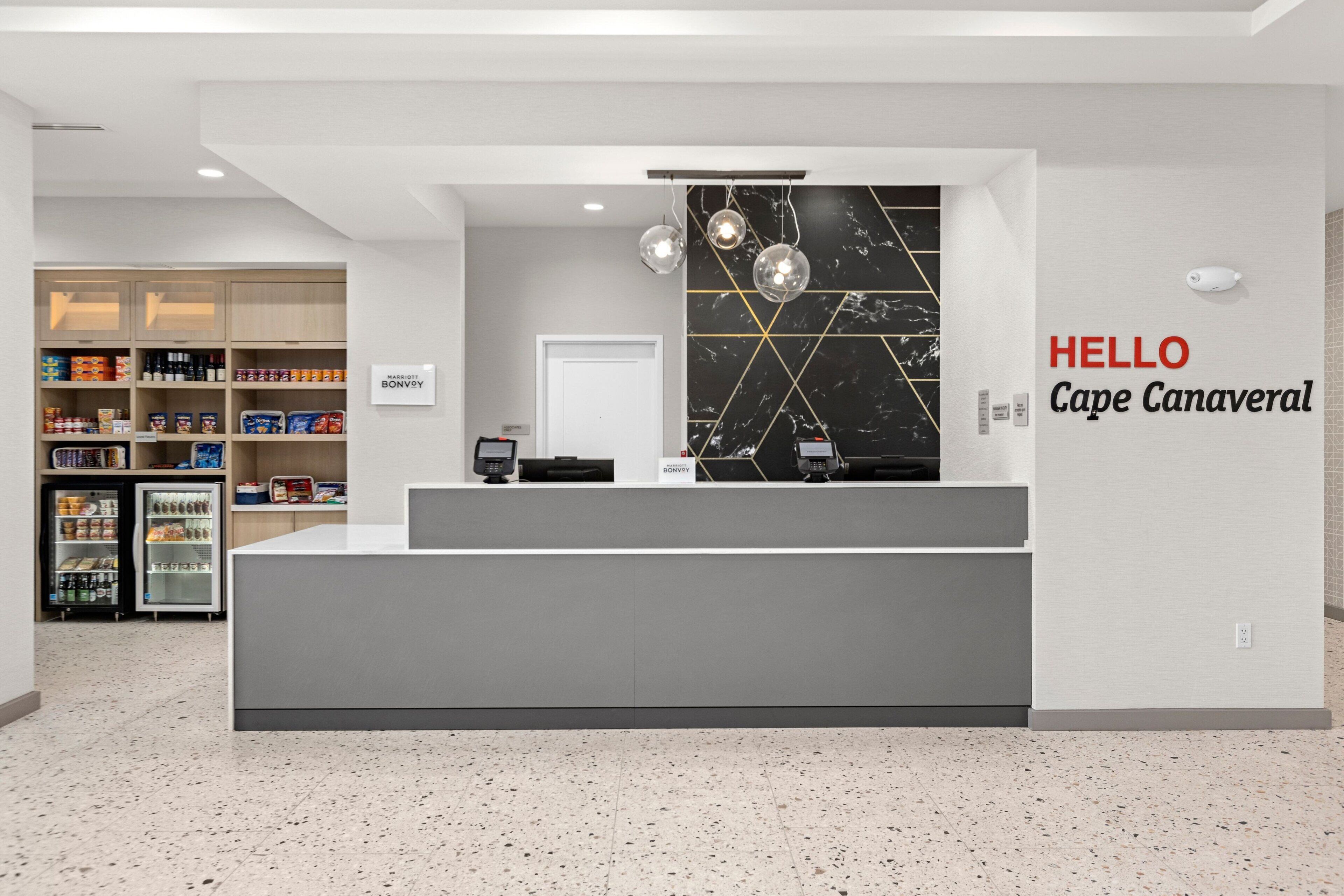 En hotellobby med modern design, inklusive en receptionsdisk, en väggmonterad skylt med texten ”HEJ Cape Canaveral” och ett kylskåp fyllt med drycker.