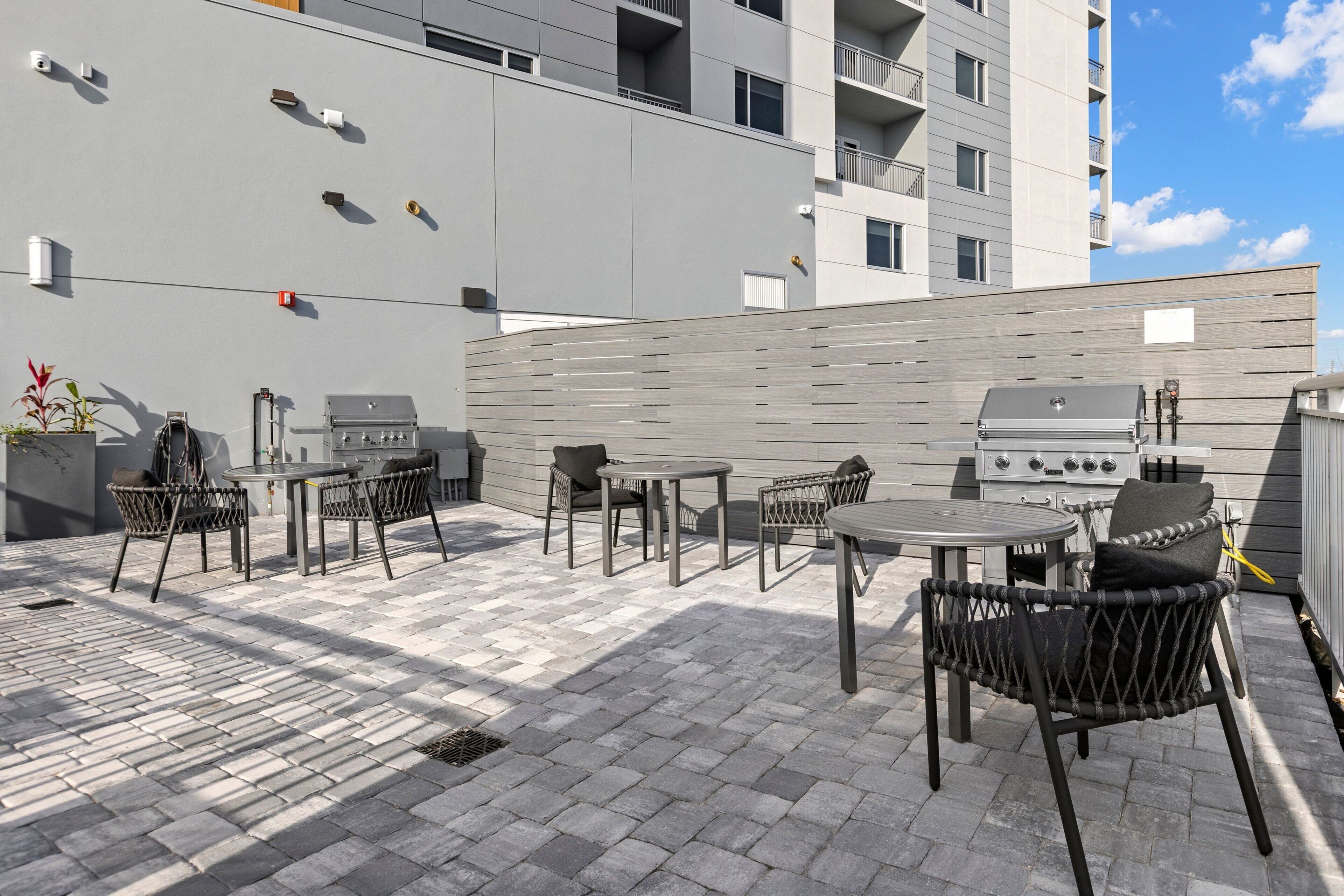 An outdoor patio with a grill, tables, and chairs, adjacent to a modern building.